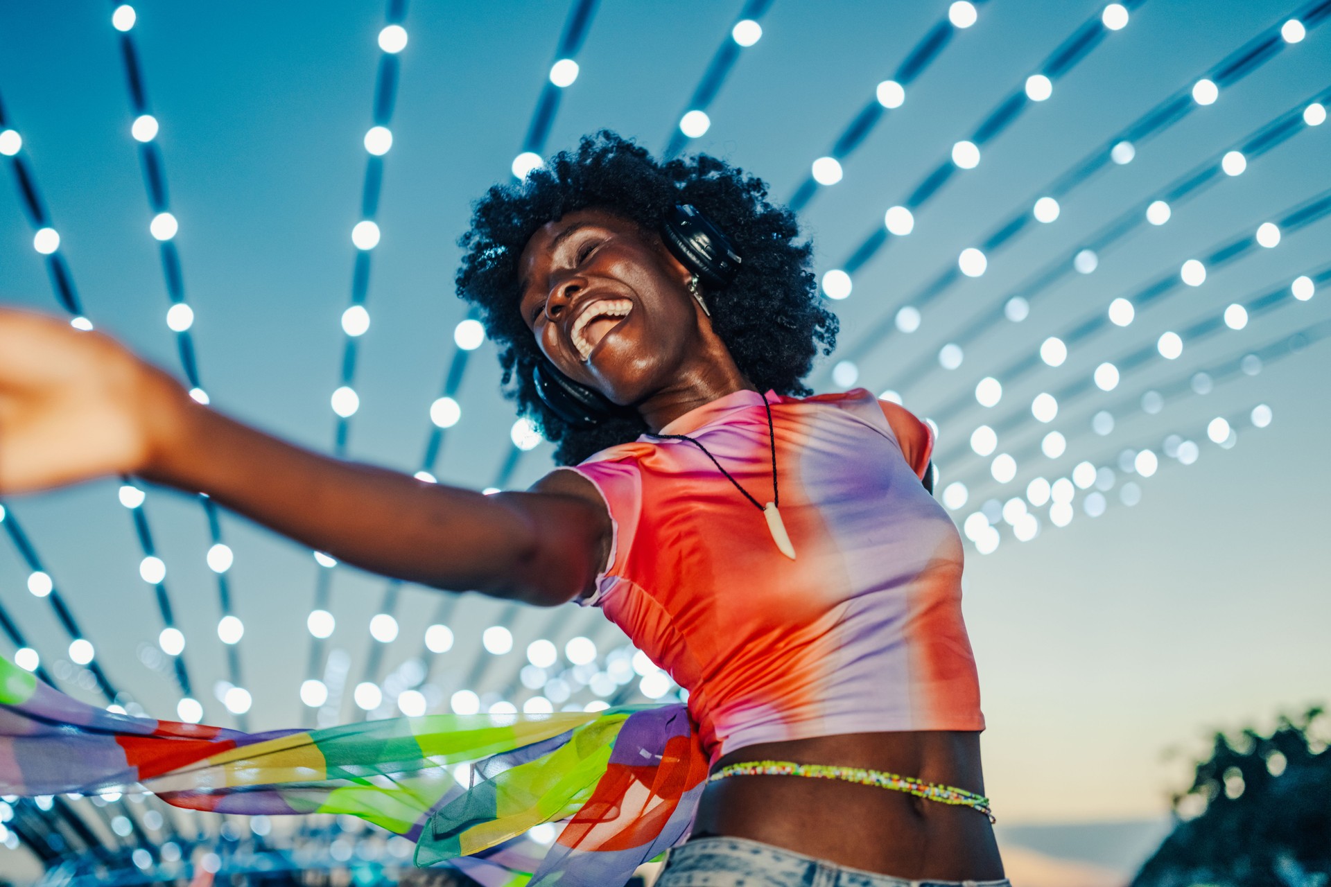 Young woman dancing with headphones and colorful scarf under string lights